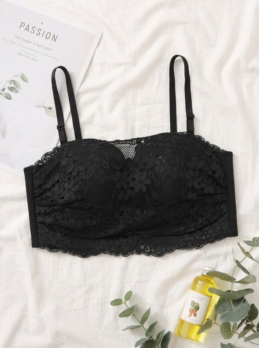 A black lace underwire bra with thin adjustable shoulder straps, laid flat on a white fabric background with decorative props including a card, eucalyptus leaves, and a small yellow bottle.