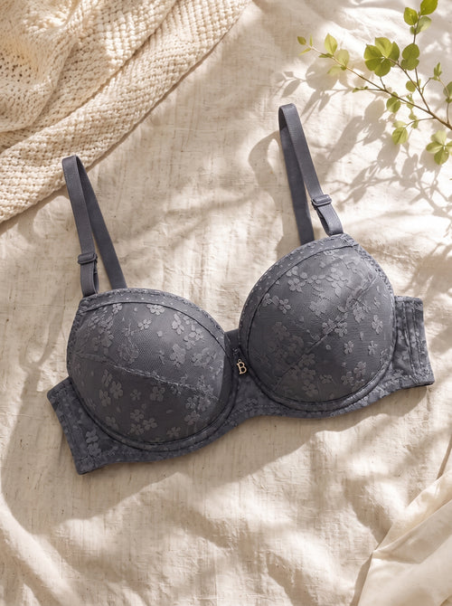 A dark gray floral lace underwire bra with adjustable straps, laid flat on a textured beige linen fabric with a knitted blanket and green leaves in the corner.