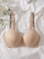A beige wireless bra laid flat on a white textured fabric background with decorative white flowers and green stems. The bra has wide adjustable shoulder straps printed with the text 'VICTORIA'S SECRET'.