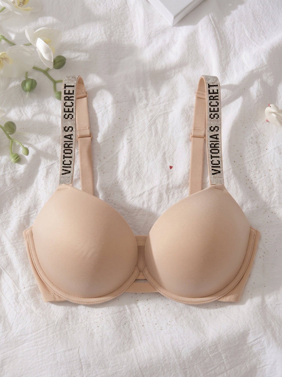 A beige wireless bra laid flat on a white textured fabric background with decorative white flowers and green stems. The bra has wide adjustable shoulder straps printed with the text 'VICTORIA'S SECRET'.