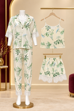A women's two-piece pajama set displayed on a mannequin, alongside matching top and shorts hanging on hangers. The pajama set features a cream-colored base with a green botanical floral print and white lace trim on the sleeve cuffs and pant hems.